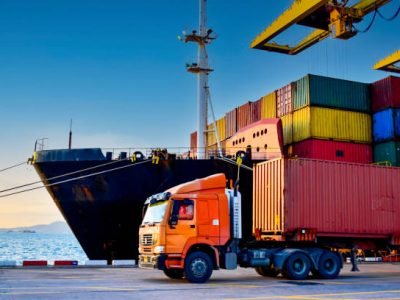 Truck carrying forty-foot container leaving port terminal with ship and quay crane on the background. Seaport operation activities, container shipping, and logistics.